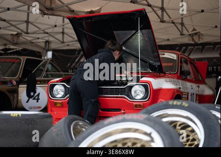 A female mechanic works on the engine of a classic red and white Ford Escort RS1600 MkI in a racing paddock with blurred tires in the foreground. Stock Photo
