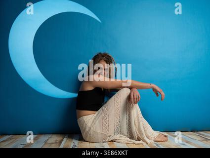 Young woman with dreadlocks sitting on wooden floor next to a blue wall with a crescent moon Stock Photo