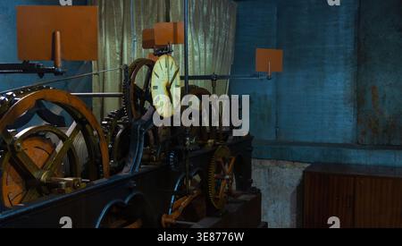 A vintage mechanical clock with a large yellow face and black hands is surrounded by rusty metal gears and cogs in a dimly lit room with blue walls, a Stock Photo