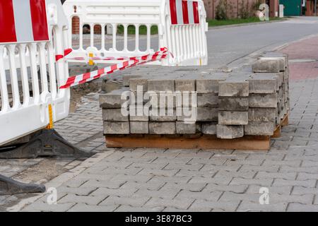 Stack of grey concrete paving blocks on wooden pallet beside white safety barriers with red tape at urban road repair site. Concept of street Stock Photo
