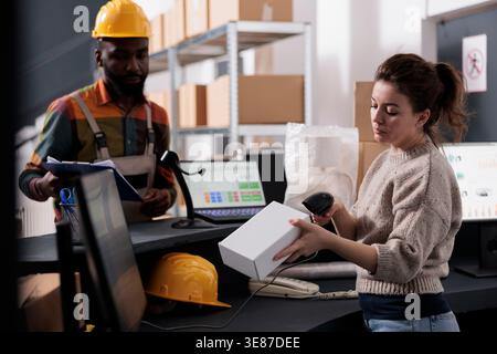 Manager scanning white carton box using store scanner, working at customer order in storehouse. African american employee with industrial overall and helmet discussing delivery details Stock Photo