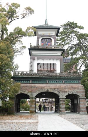 Traditional gate with stone arches and green roof. Tall tower stands behind, framed by trees. Two people walk through archway, distant and calm. Path Stock Photo