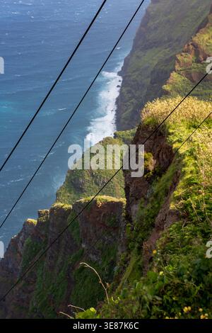 Steep emerald cliffs meet the Atlantic as waves foam on a pebble shore. Three slanted cable lines cross the scene in soft afternoon light on Madeira's Stock Photo