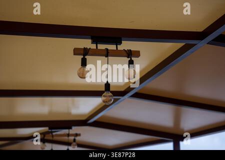 Ceiling fixture with three round bulbs on a wooden crossbar, framed by dark metal beams and matte beige panels, indoors at a Madeira island cafe or re Stock Photo