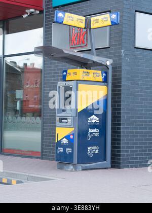 Automated cash machine stands outside shop. Machine features vibrant yellow and blue colors. Brick wall and glass storefront form backdrop. No people Stock Photo
