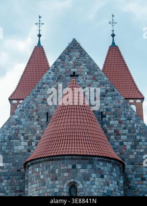 Stone church with red-tiled spires under soft clouds. Grey stones form ancient, sturdy structure. Weathered rooflines reach toward the sky. Cross mark Stock Photo