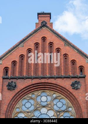 Red brick facade with arched stained glass window. Decorative brickwork features vertical niches and gable. Black wrought iron embellishments flank th Stock Photo