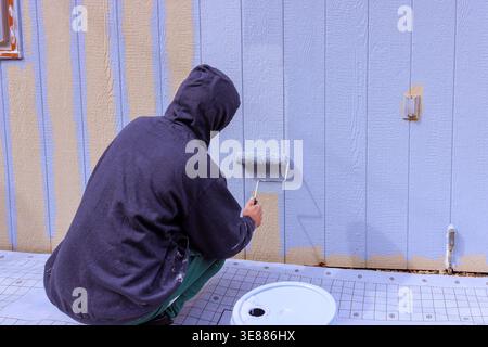 Someone is applying paint to siding panel wall using roller while sitting on ground in construction setting. Stock Photo