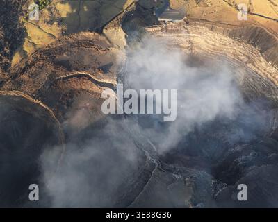 Thick smoke billows from a volcanic crater in a rural area during the daytime. The surrounding land shows signs of volcanic activity. Stock Photo