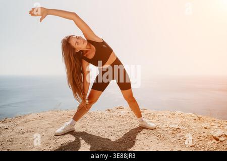 Yoga stretching cliffside young woman performs outdoor fitness workout on a rocky edge overlooking the bright blue sea Stock Photo