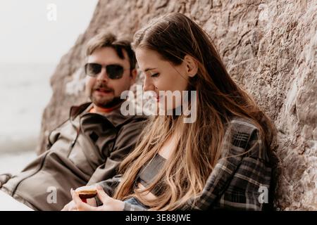 Couple picnic beach woman in plaid shirt eats snack with her boyfriend sitting by rock wall during afternoon outdoors Stock Photo