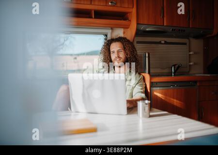 Young man working remotely from caravan. Man using camper as homeoffice, working on notebook. Stock Photo