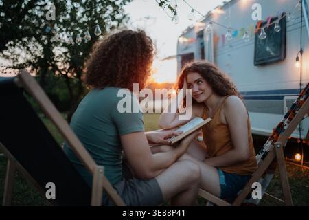 Couple having romantic camping trip in nature. Man reading book to girlfriend. Caravan traveling for young people. Stock Photo