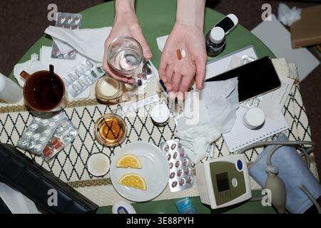 Top view of adult hands holding pills and water above table with medicine, tissues, thermometer, blood pressure monitor, signaling home treatment. Useful for healthcare ads, cold flu content Stock Photo