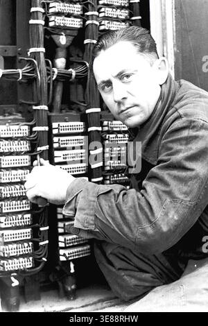 Sloviansk, 1956. View inside a local Automatic Telephone Exchange (ATS). A focused male technician wearing a dark work uniform is carefully inspecting and repairing a complex communication switchboard. He is surrounded by an intricate maze of thick electrical cables, wires, and numerous connection terminals, looking directly at the camera with a serious expression. This striking black and white historical archive photograph captures the specialized labor, technological progress, and working professionals in a peaceful Donbas. Stock Photo