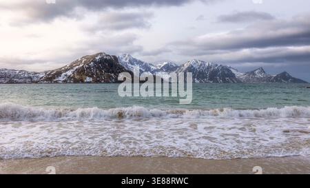 Waves crash gently on soft sands while snow-capped mountains rise dramatically under a moody sky at Haukland Beach, a serene spot in Lofoten, Norway. Stock Photo