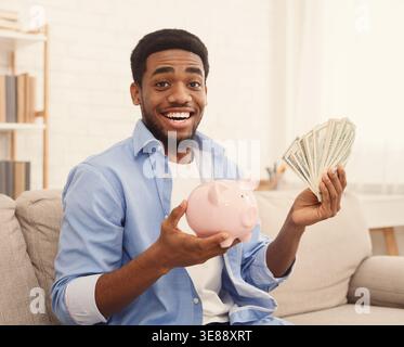 Excited african-american guy holding piggybank and dollar bills Stock Photo