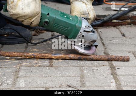 Close-up of a worker using an angle grinder to cut a rusty rebar on a paved surface, producing bright sparks. Protective gloves and power tool convey Stock Photo