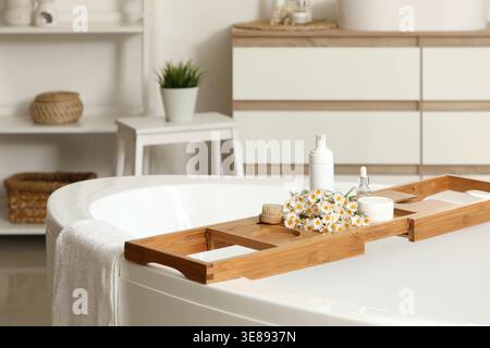 Wooden board with artificial flowers and bathing accessories on bathtub in bathroom, closeup Stock Photo