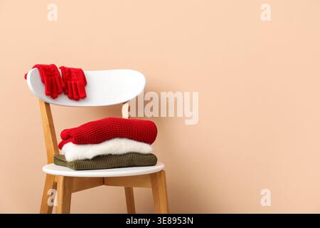 Stack of folded sweaters with gloves on chair against beige background Stock Photo