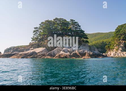 Green pine trees cover a dramatic rocky island shoreline extending into deep blue ocean water on a clear sunny day Stock Photo