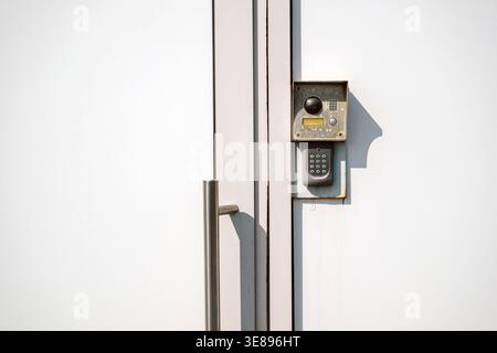 Architectural detail of a weathered metal intercom keypad and doorbell system mounted on a smooth white door on bright sunny day Stock Photo