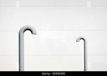 Architectural close-up of two grey metal pipes with smooth curved J-shaped tops mounted on a bright white horizontal-ribbed industrial wall. Selective Stock Photo