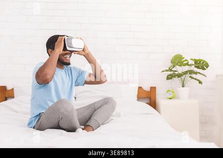 Man Using Virtual Reality Headset in Bedroom While Sitting on Bed and Smiling During Leisure Time Stock Photo