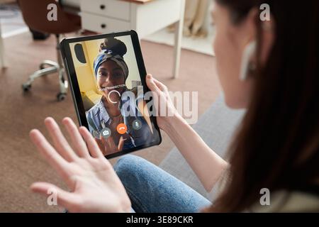 Over shoulder shot of young adult Caucasian woman video calling African American woman on tablet, gesturing during conversation. Ideal for remote communication, social connection, digital lifestyle Stock Photo