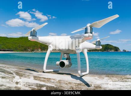 Modern white camera drone positioned on coastal surface overlooking beautiful blue ocean waters and green hills on a sunny day Stock Photo