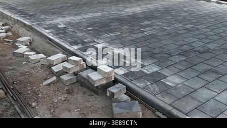 Paved surface with gray concrete blocks partially laid on construction site, scattered loose blocks and gravel visible along the edge of the pavement Stock Photo