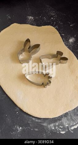Metal cookie cutters in the shapes of a bunny, a chick, and a carrot placed on rolled-out dough on a dark countertop ready for baking Stock Photo