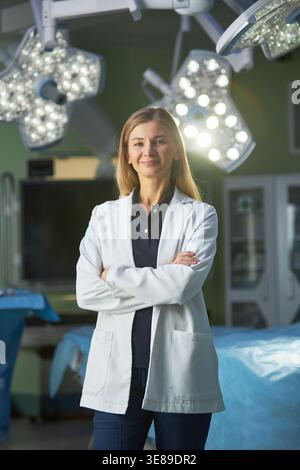 Portrait of Caucasian young adult female surgeon standing with crossed arms in operating room, conveying medical expertise and leadership. Useful for healthcare staffing, hospital branding, vertical Stock Photo