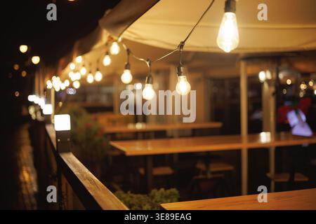 Garlands of light bulbs glow against the backdrop of tables at a summer cafe. Warm, cozy cafe lighting. Cafe terrace. Stock Photo