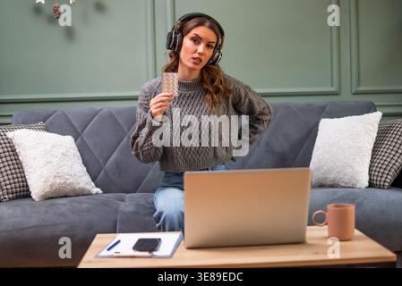 Young woman is sitting on bed in living room and having online doctor's appointment using laptop. Young woman taking medication while working on lapto Stock Photo