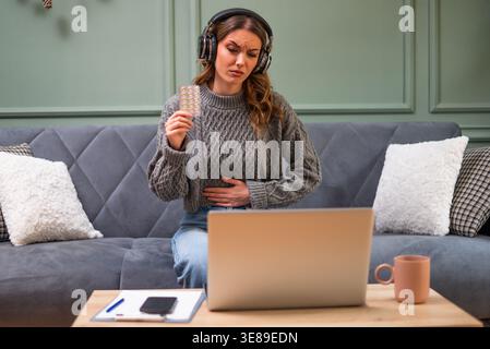 Young woman is sitting on bed in living room and having online doctor's appointment using laptop. Young woman taking medication while working on lapto Stock Photo