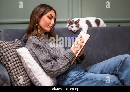 Cheerful cozy dressed woman sitting on sofa and writing diary. Her cat is lying beside her. Stock Photo