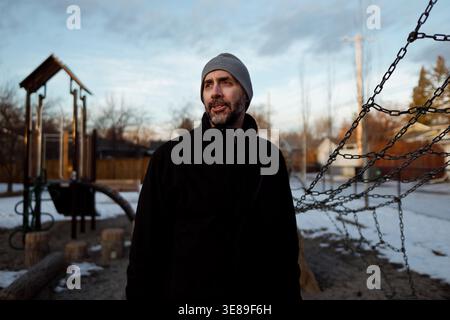 Middle aged man in a beanie standing at a snowy playground at dusk Stock Photo