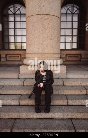 Stylish woman in leather jacket sitting on stone steps in city Stock Photo
