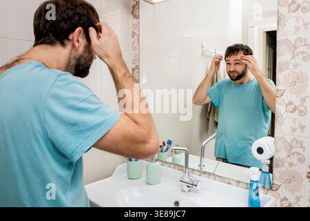 Sleepy man looking at himself in mirror during early morning routine Stock Photo