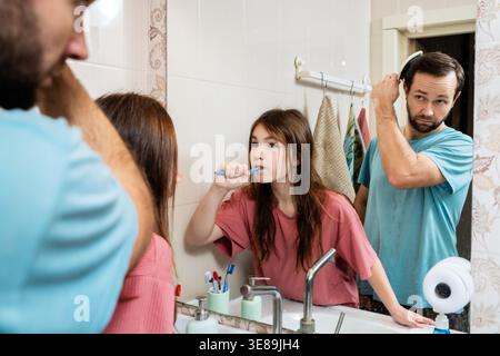 Father brushing hair and daughter brushing teeth in front of mirror. Stock Photo
