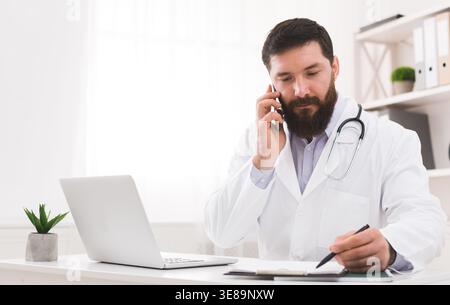 Doctor on the Phone While Taking Notes at the Desk in His Office During Daytime Stock Photo