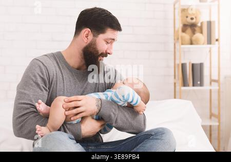 Father Holds Baby While Sitting on Bed in Bright Room With Teddy Bear on Shelf Stock Photo