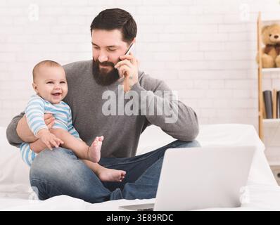 Father Sits on Bed With Baby While Talking on Phone and Using Laptop in a Home Setting Stock Photo