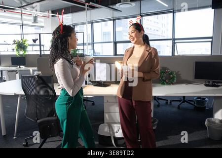 Diverse female coworkers chatting, holding coffee cups, wearing work attire and headbands in office. Colleagues, workspace, collaboration, holiday, wreath, monitors, plants Stock Photo