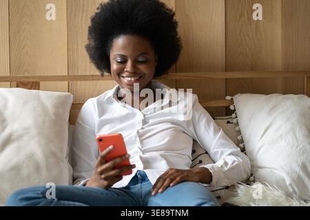 Cheerful Afro American millennial young woman lying on bed at home, resting, using mobile phone, typing message her boyfriend, taking a break from work. Black female chatting in social media. Stock Photo