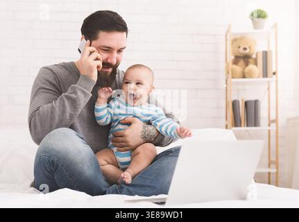 Father Talks on Phone While Holding Baby in His Lap and Using Laptop at Home in Cozy Setting Stock Photo