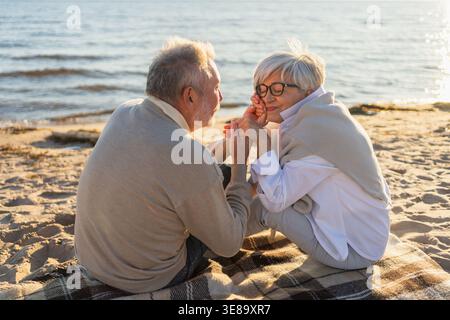 Senior mature couple sitting on beach holding hands enjoying outdoor recreation. Old husband wife touching hands with tenderness love. Grandmother grandfather together. Family moment of love Stock Photo