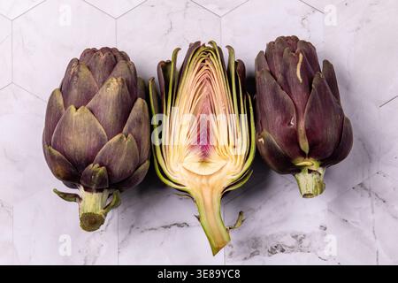 Three artichokes placed on a marble surface, one sliced. Stock Photo