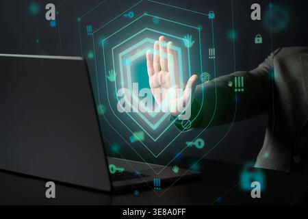 Businessman raises his hand againt a laptop behind a glowing digital green shield hologram as floating icons suggest data security privacy controlled Stock Photo